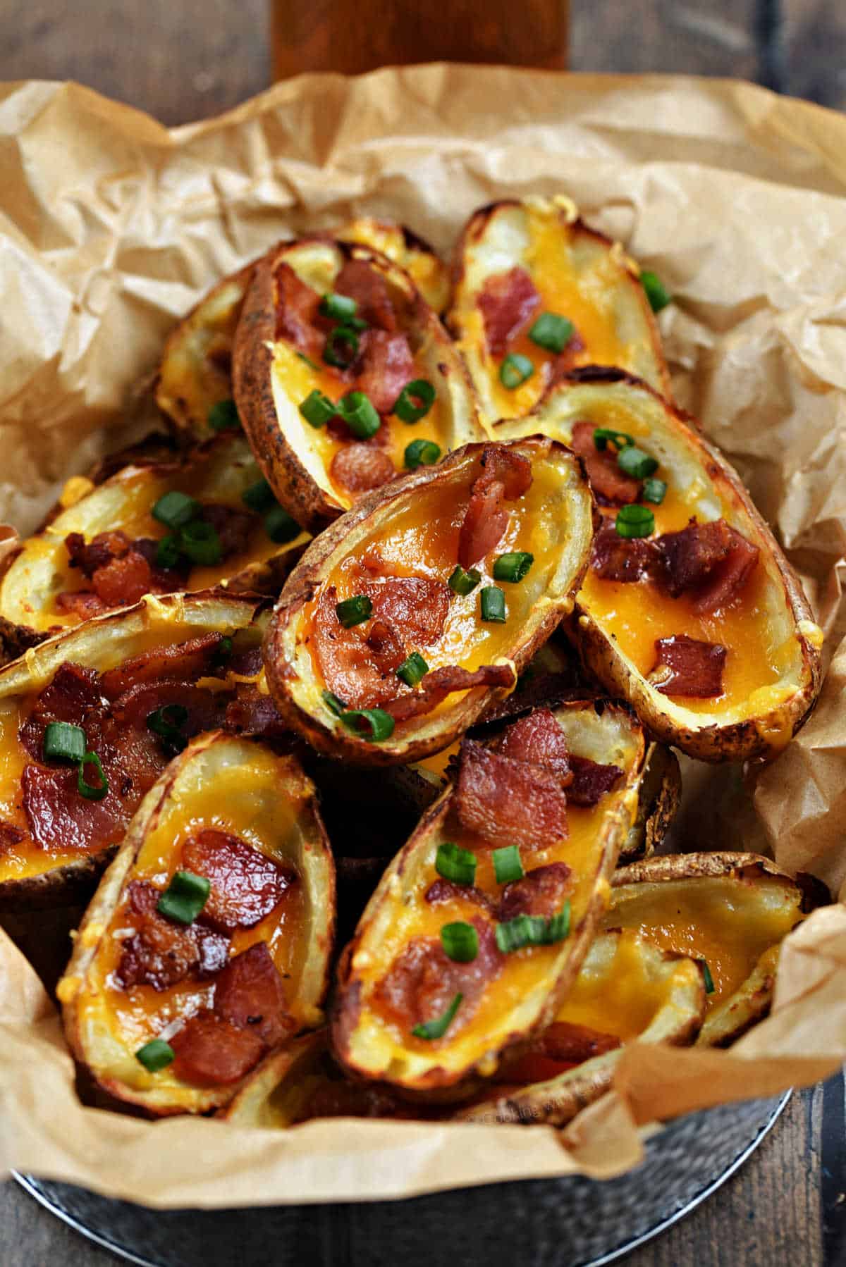 Crispy potato skins topped with melted cheddar and bacon pieces stacked in a parchment paper lined basket.
