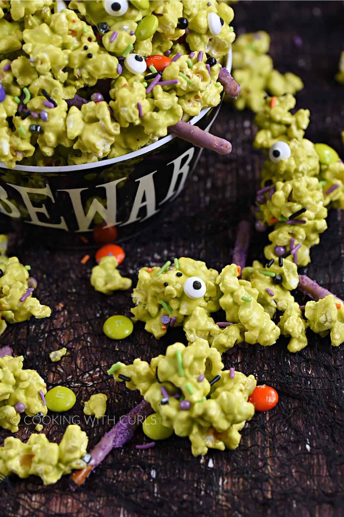 Green candy coated popcorn with candy eyes, sprinkles, purple pretzel sticks, and candy coated chocolates overflowing a bowl onto a wood surface. 