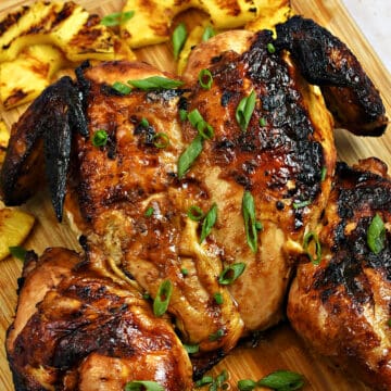 A whole grilled chicken on a wood board surrounded by grilled pineapple slices.